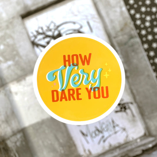 A yellow sticker with orange-red, light blue, and white text that says How Very Dare You. The word "dare" is written in script and the rest of the words are in blocky capital letters. The sticker sits above a blurred black and white background.