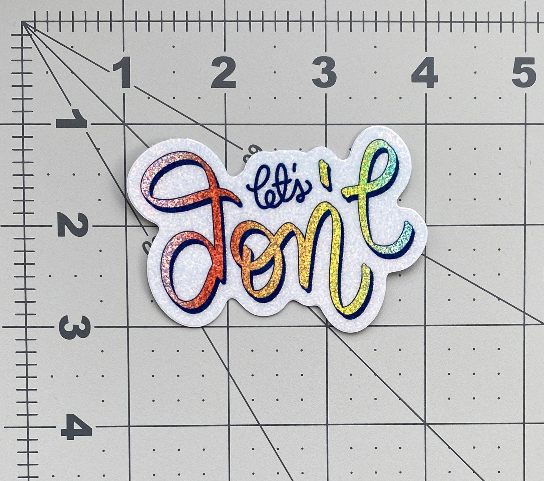 A rainbow glitter sticker that says "let's don't" in calligraphy and sits on a ruled background to show it's size.