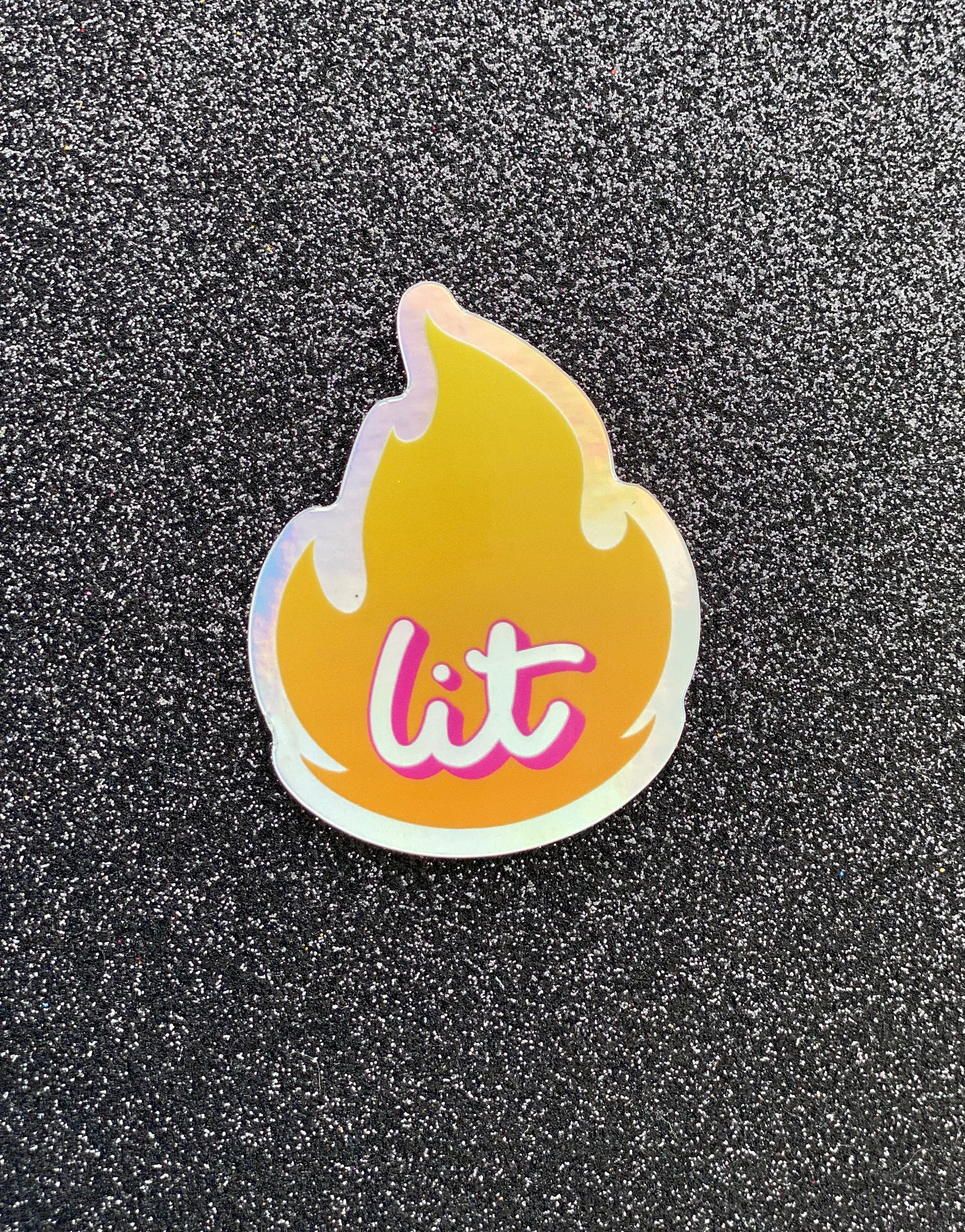 Two holographic stickers, one larger, one smaller. The sticker is an illustration of the "fire" emoji and inside is the word "lit" in script. The sticker sits on a black glitter background.