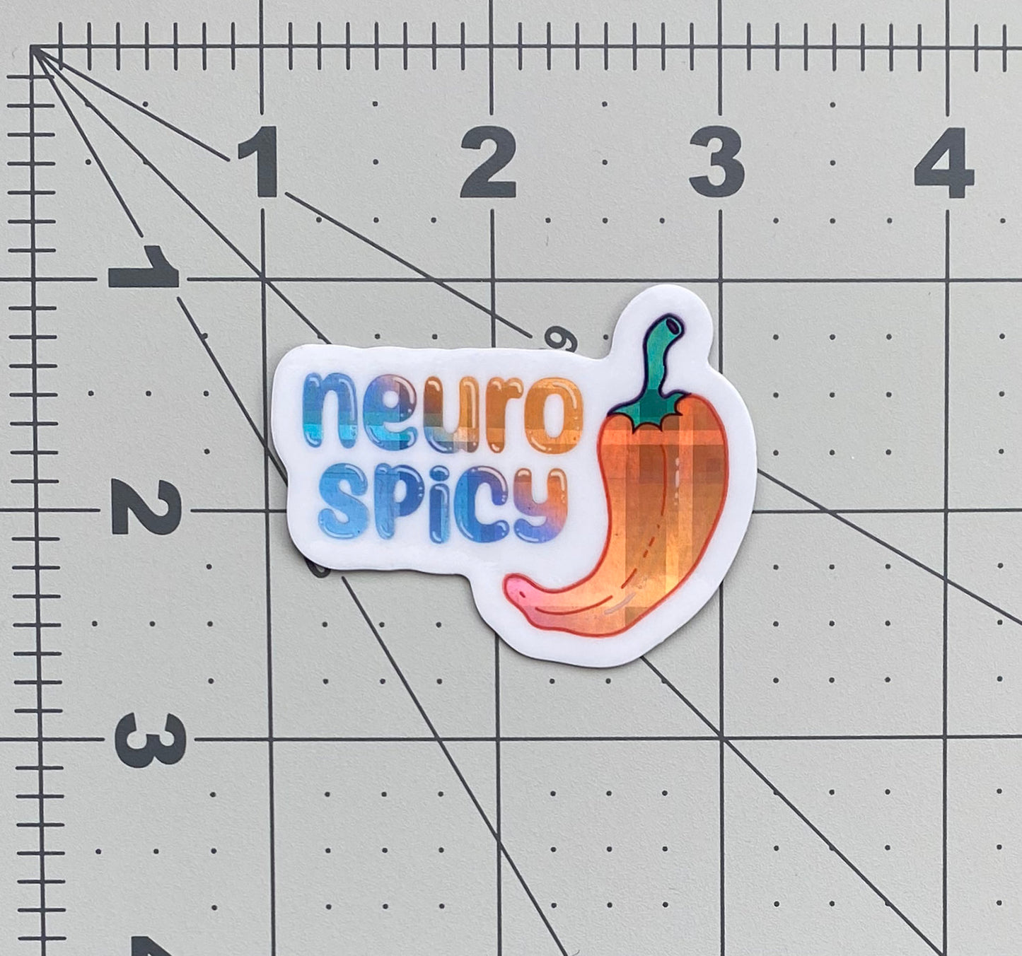 A holographic vinyl sticker with an illustration of a pepper and the words "neuro spicy". The holographic material is a blue to purple to orange gradient pattern with a plaid effect. The sticker sits on a ruled background to show its size.