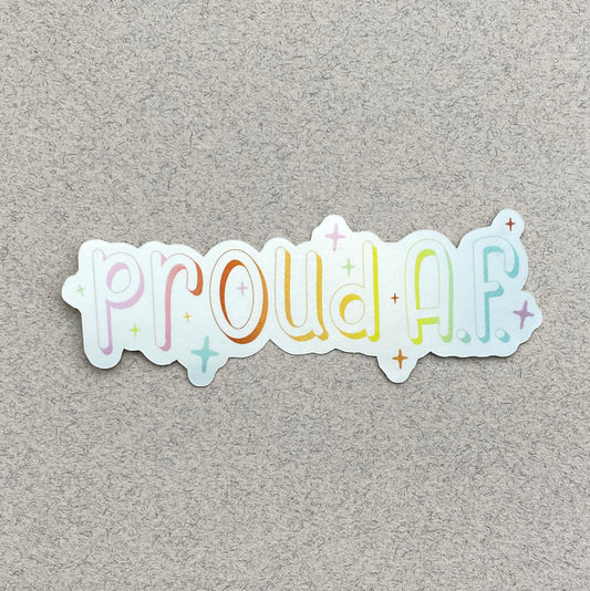A metallic silver sticker against a varigated gray paper background. The sticker says "proud A.F." in a pastel rainbow gradient and is surrounded by sparkles.