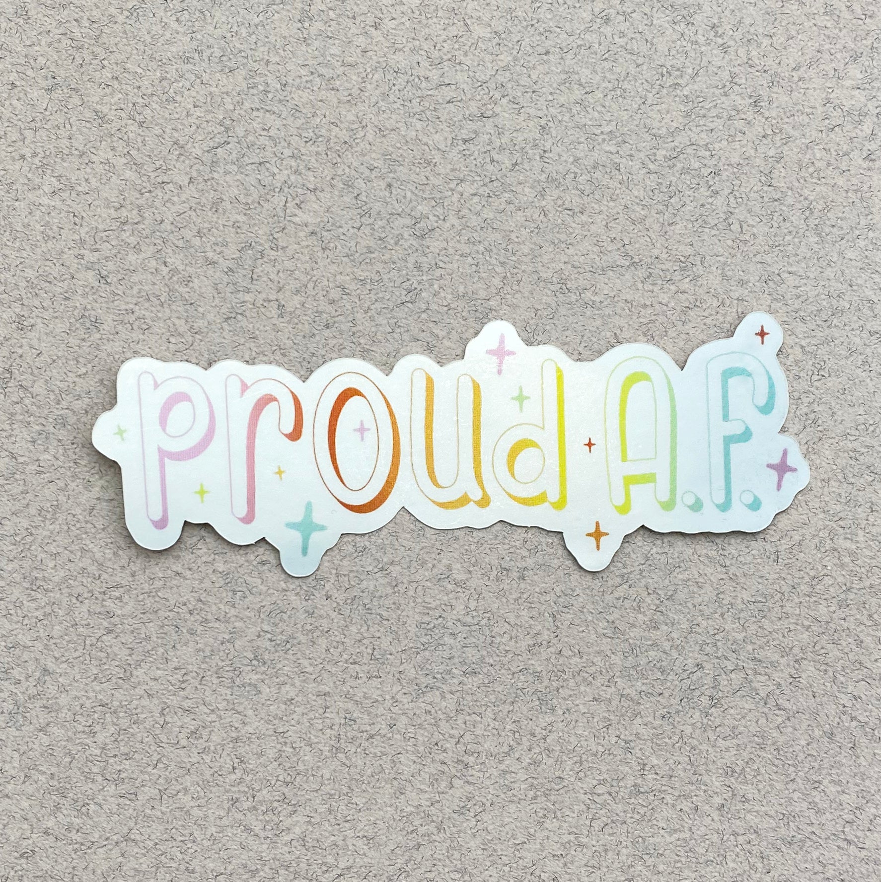 A metallic silver sticker against a varigated gray paper background. The sticker says "proud A.F." in a pastel rainbow gradient and is surrounded by sparkles.