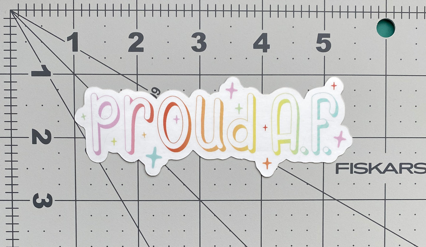 A metallic silver sticker against a ruled background to show its size. The sticker says "proud A.F." in a pastel rainbow gradient and is surrounded by sparkles.