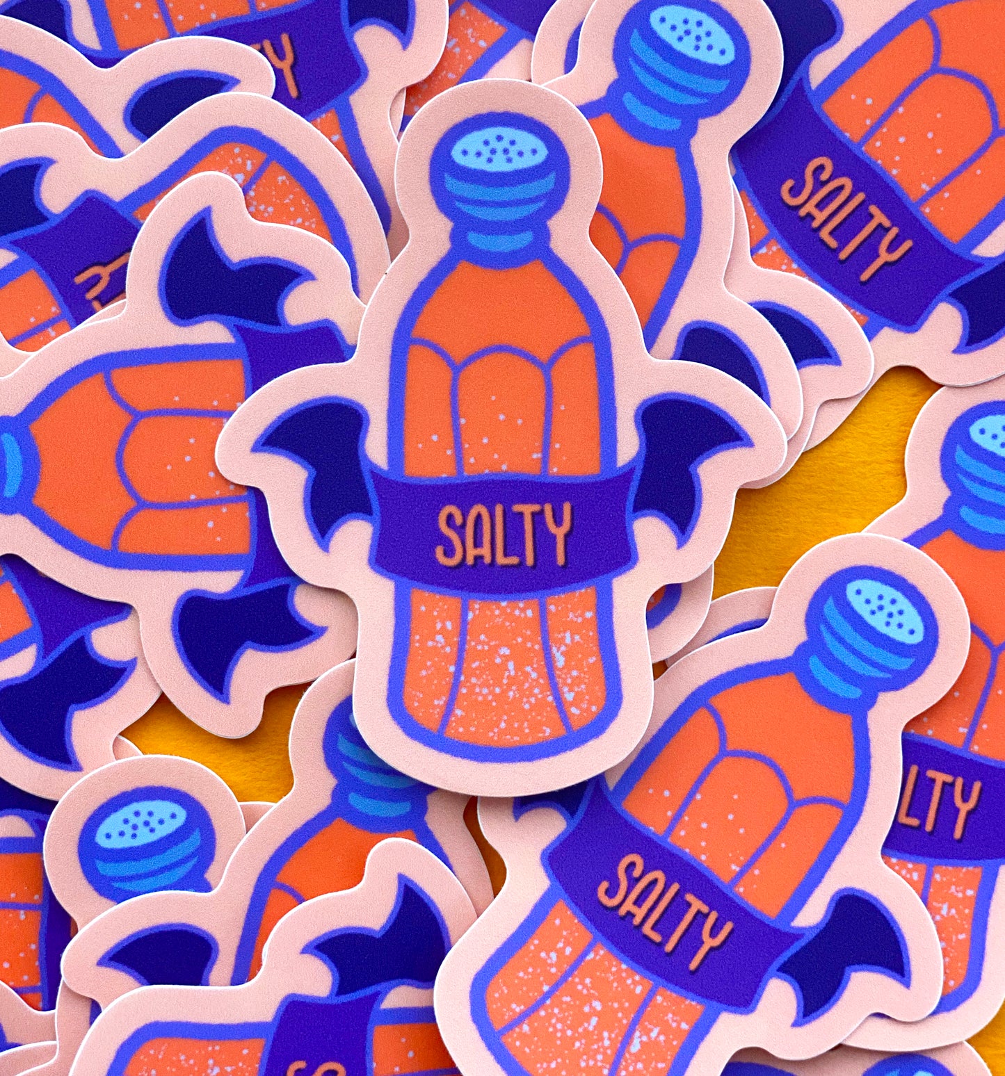 A pile of peach vinyl stickers with an illustration of a salt shaker and a banner that says "Salty" against a bright orange background.