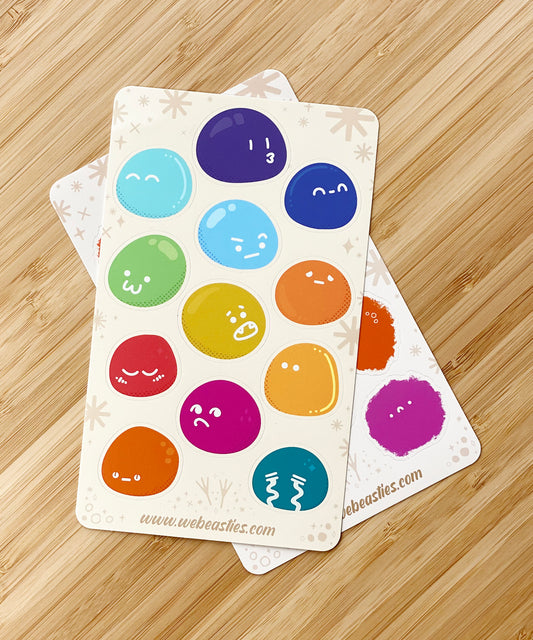 A sticker sheet with 12 slimes of different colors with different expressions. The Slimes sticker sheets sit on top of the Fluffies sticker sheet, both on a bamboo table top.