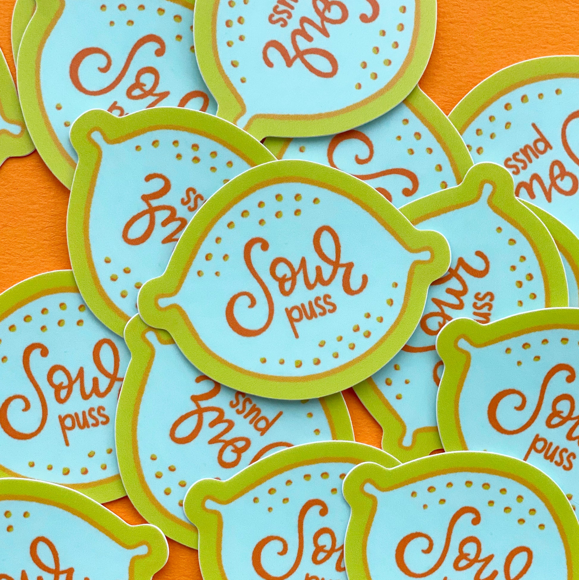 A pile of vinyl stickers that feather an illustration of a blue lemon and the words "Sour puss" written on it. They sit on a bright orange paper background.