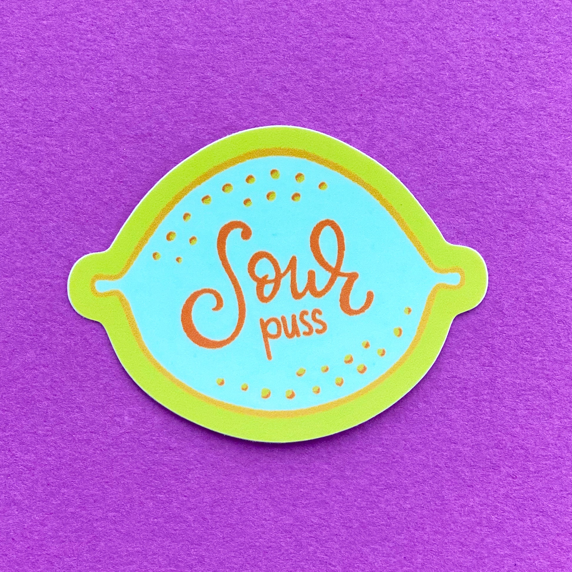 A vinyl sticker with an illustration of a blue lemon and the words "Sour puss" written on it. The sticker has a yellow border and sits on a birght purple paper background.