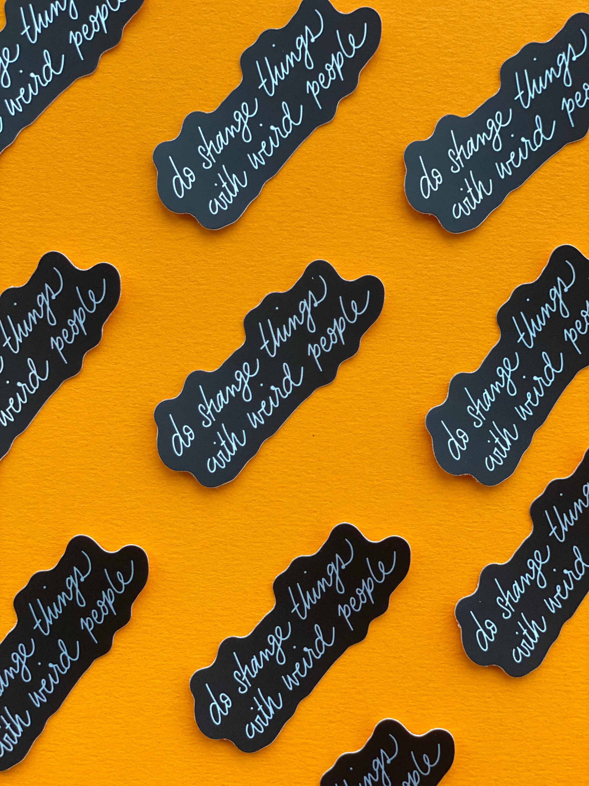 A handful of black stickers with silver text that says "do strange things with weird people" on a bright orange background.