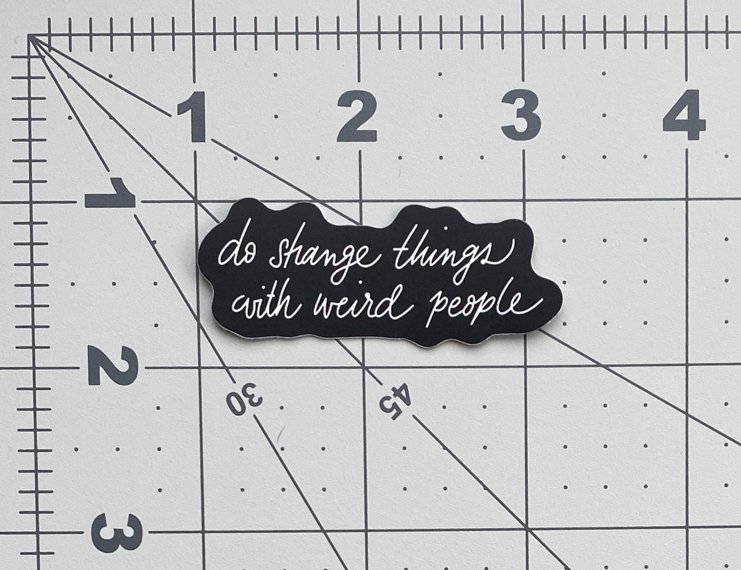 A black sticker with silver lettering that says "do strange things with weird people" against a ruled background to show size.