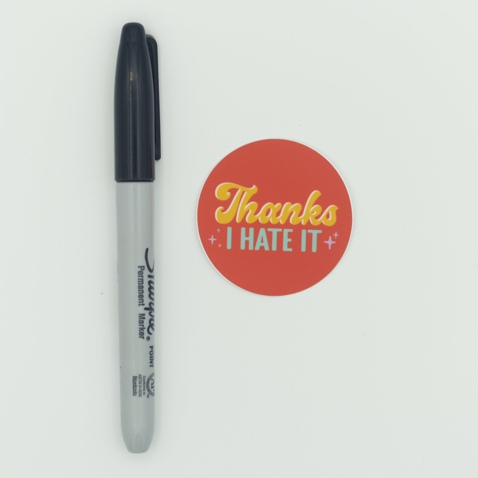 An orange sticker with the words "Thanks I hate it" in yellow and light blue text next to a Sharpie for scale.