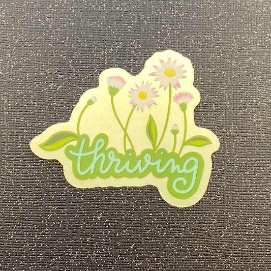 A metallic sticker with the word "thriving" growing white, pink, and yellow flowers on a pale green metallic background. Sticker sticks on black glitter paper.