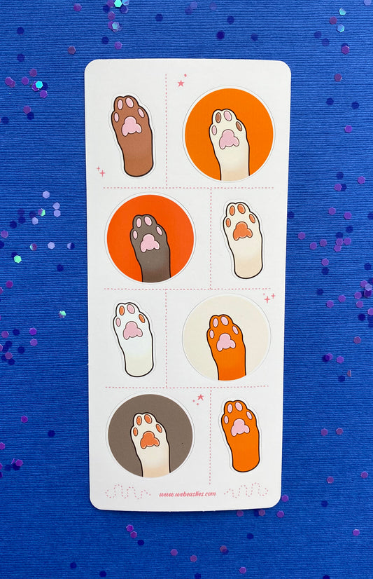 A sticker sheet featuring 8 illustrations of cat's paws and toe pads. The paws are light brown, dark brown, white, and off white. The toe pads are pink, brown, or both. Four paws sit against orange, dark orange, cream or light brown backgrounds and the others are an outline of the paw with no background. The sticker sheet sits against a blue paper background scattered with chunky blue and purple glitter. 