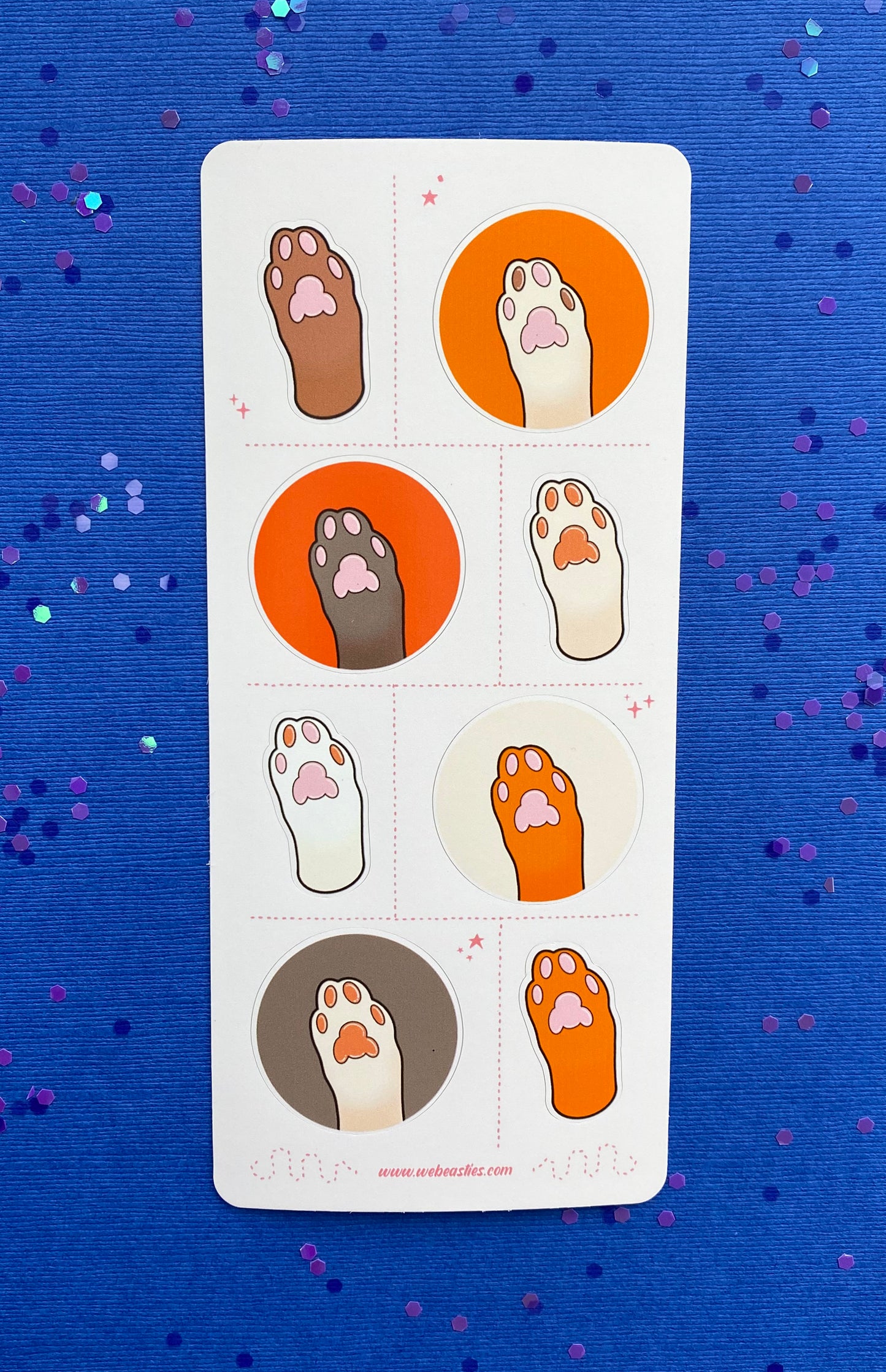 A sticker sheet featuring 8 illustrations of cat's paws and toe pads. The paws are light brown, dark brown, white, and off white. The toe pads are pink, brown, or both. Four paws sit against orange, dark orange, cream or light brown backgrounds and the others are an outline of the paw with no background. The sticker sheet sits against a blue paper background scattered with chunky blue and purple glitter. 