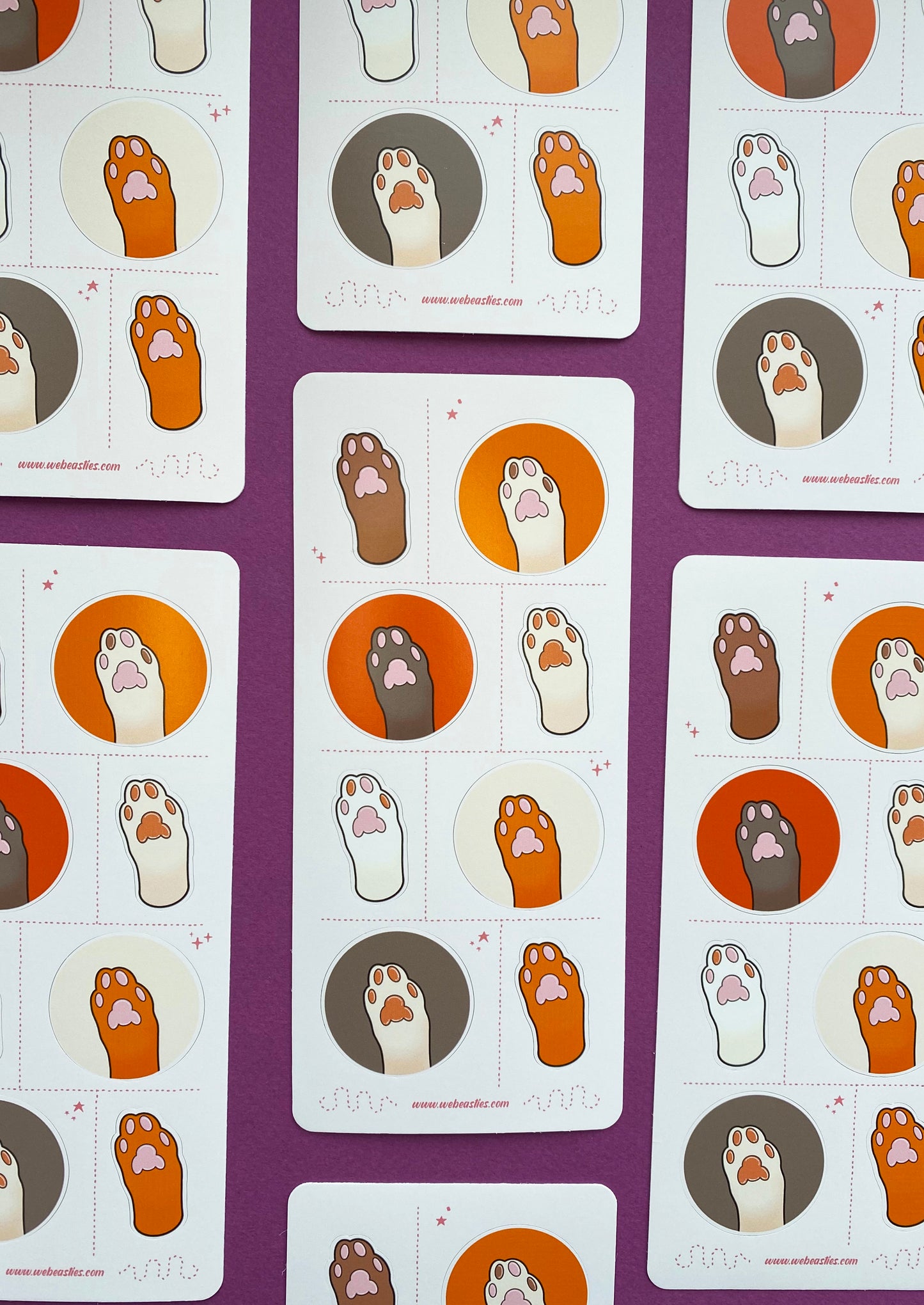 A collection of sticker sheets featuring 8 illustrations of cat's paws and toe pads. The paws are light brown, dark brown, white, and off white. The toe pads are pink, brown, or both. Four paws sit against orange, dark orange, cream or light brown backgrounds and the others are an outline of the paw with no background. The sticker sheet sits against a purple background.