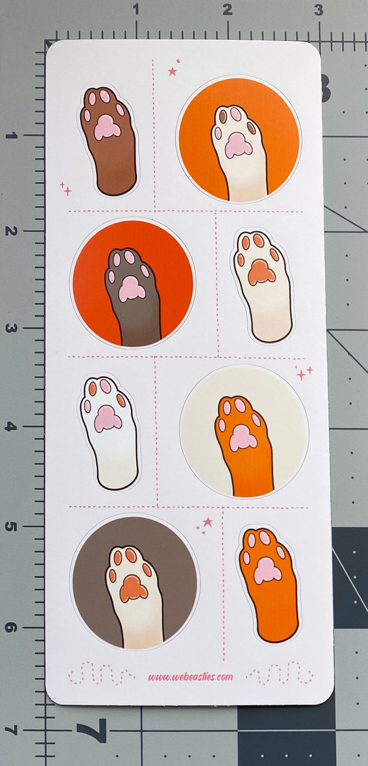 A sticker sheet featuring 8 illustrations of cat's paws and toe pads. The paws are light brown, dark brown, white, and off white. The toe pads are pink, brown, or both. Four paws sit against orange, dark orange, cream or light brown backgrounds and the others are an outline of the paw with no background. The sticker sheet sits against a ruled background to show size.