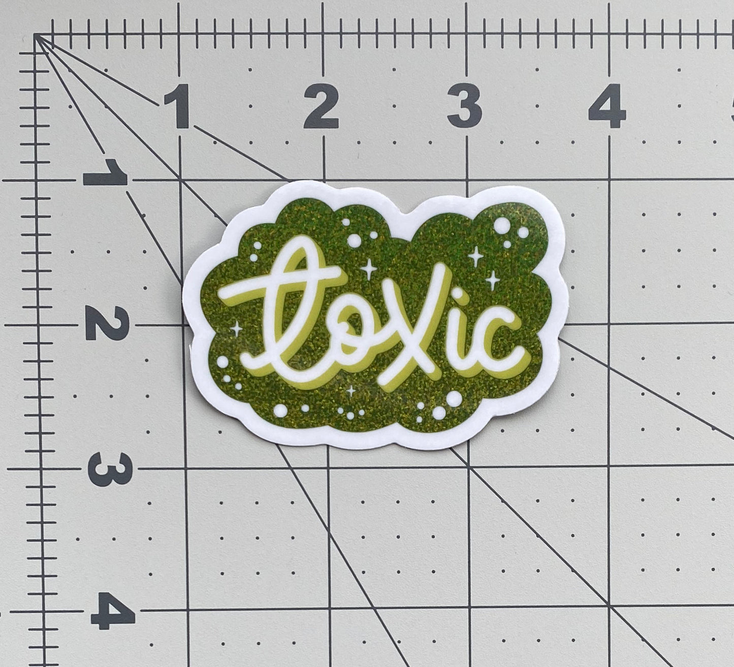 A pile of green glitter stickers that say "Toxic" in script. The sticker sits on a ruled background to show size.